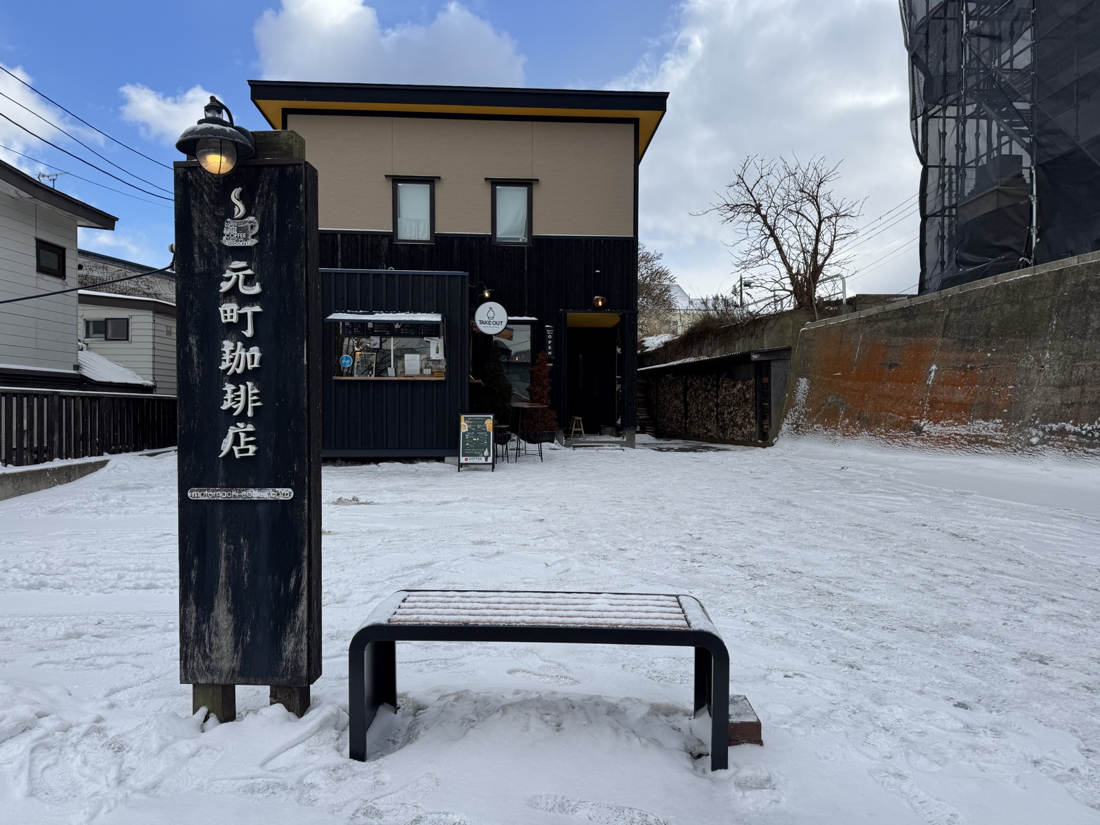 A snow-covered street with a wooden coffee-shop sign and a lone bench