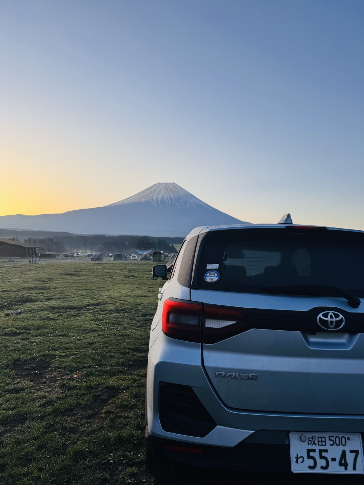 Mount Fuji at dawn seen over a campsite, with a parked car in the foreground