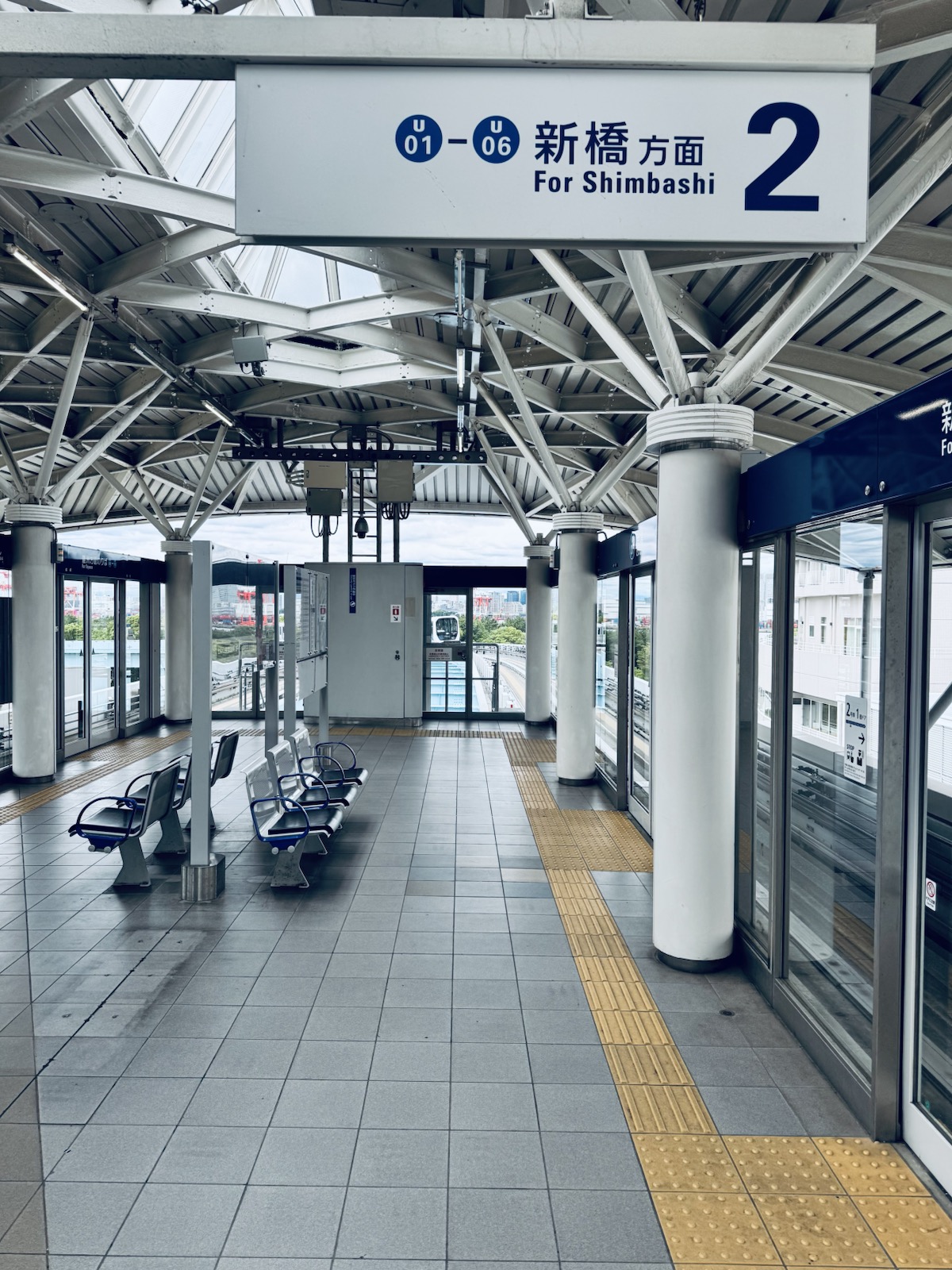 An empty elevated train platform with glass doors and blue seats, a sign reading For Shimbashi 2